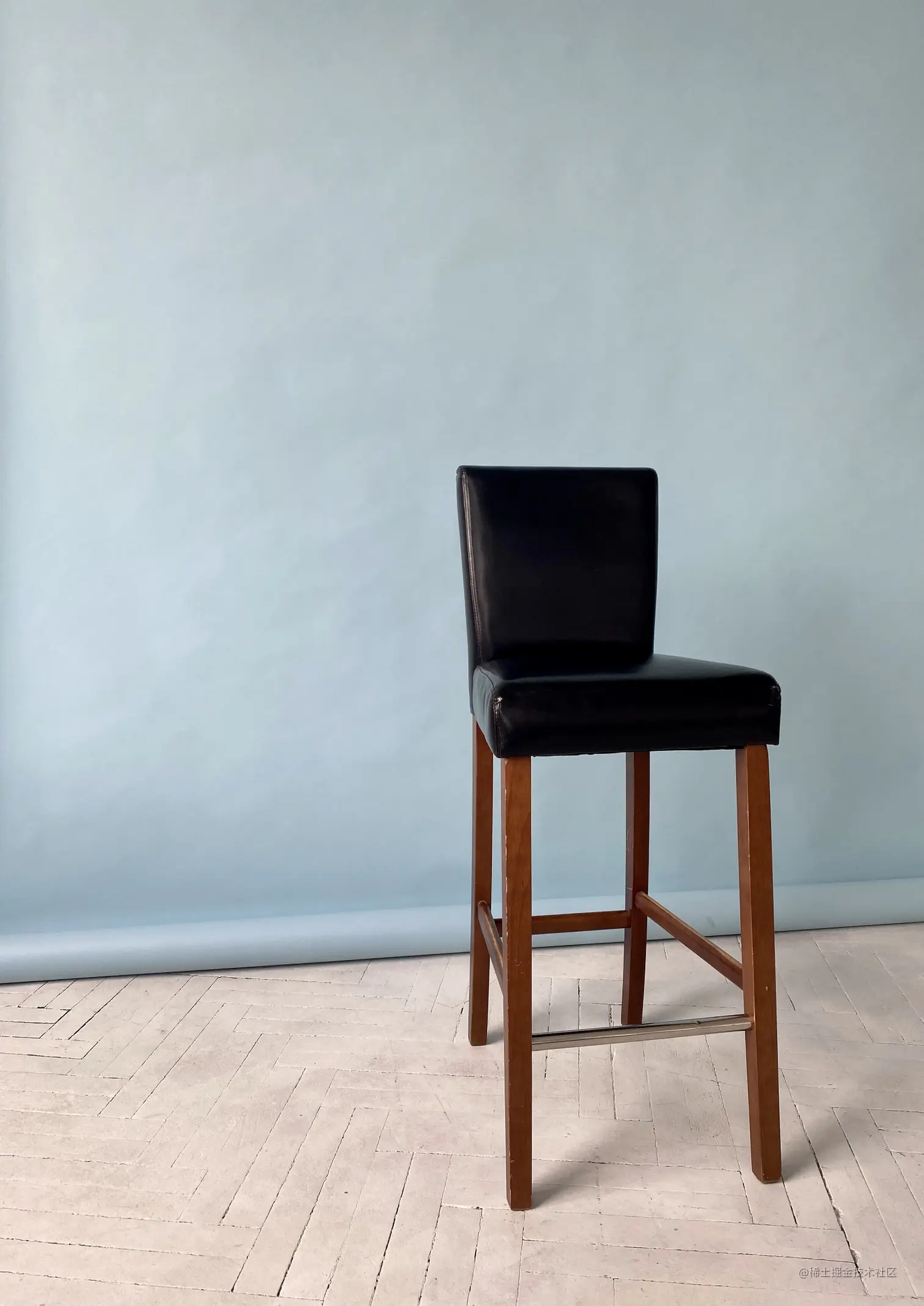 A tall black leather chair with a back and four brown legs against a light blue background.